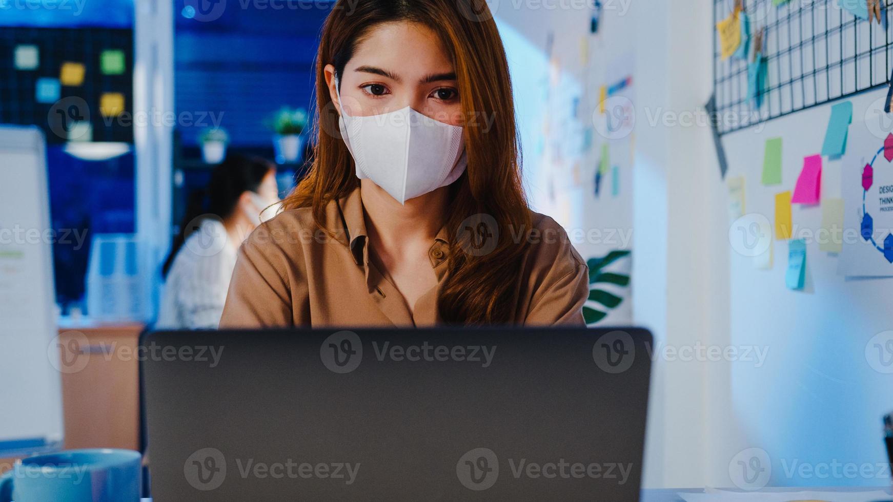 Fröhliche Geschäftsfrau in Asien, die eine medizinische Gesichtsmaske für soziale Distanzierung in einer neuen normalen Situation zur Virenprävention trägt, während sie in der Büronacht den Laptop bei der Arbeit verwendet. Leben und Arbeiten nach dem Coronavirus. foto