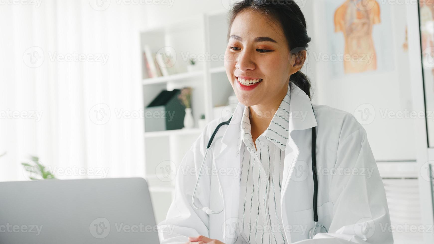 junge asiatische Ärztin in weißer medizinischer Uniform mit Stethoskop mit Computer-Laptop, die Videokonferenz mit Patienten am Schreibtisch in einer Klinik oder einem Krankenhaus spricht. Beratungs- und Therapiekonzept. foto