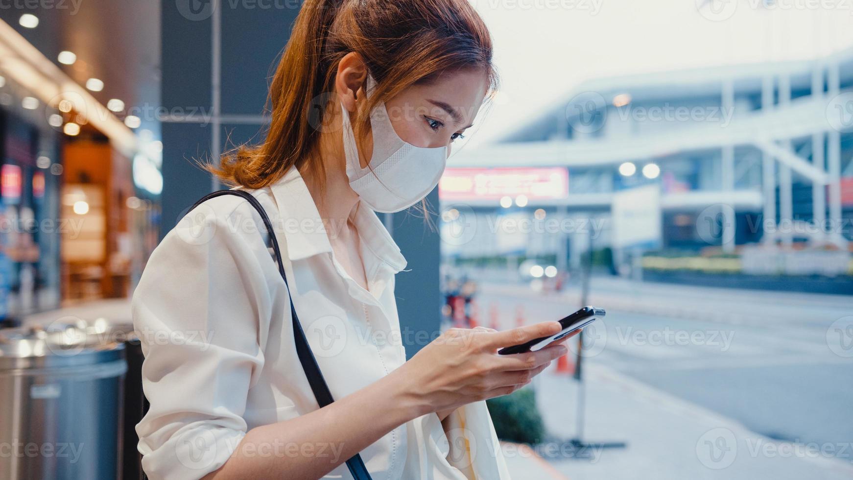 junge asiatische Geschäftsfrau in modischer Bürokleidung mit medizinischer Gesichtsmaske mit Smartphone, die Textnachrichten schreibt, während sie nachts in der urbanen modernen Stadt im Freien sitzt. Business-on-Go-Konzept. foto