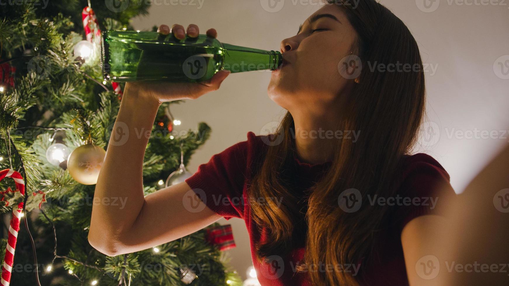 junge asiatische frau, die bier trinkt und spaß hat, fröhliche nachtparty, videoanruf, gespräch mit paar, weihnachtsbaum, der mit ornamenten im wohnzimmer zu hause geschmückt ist. Weihnachtsnacht und Neujahrsfest. foto