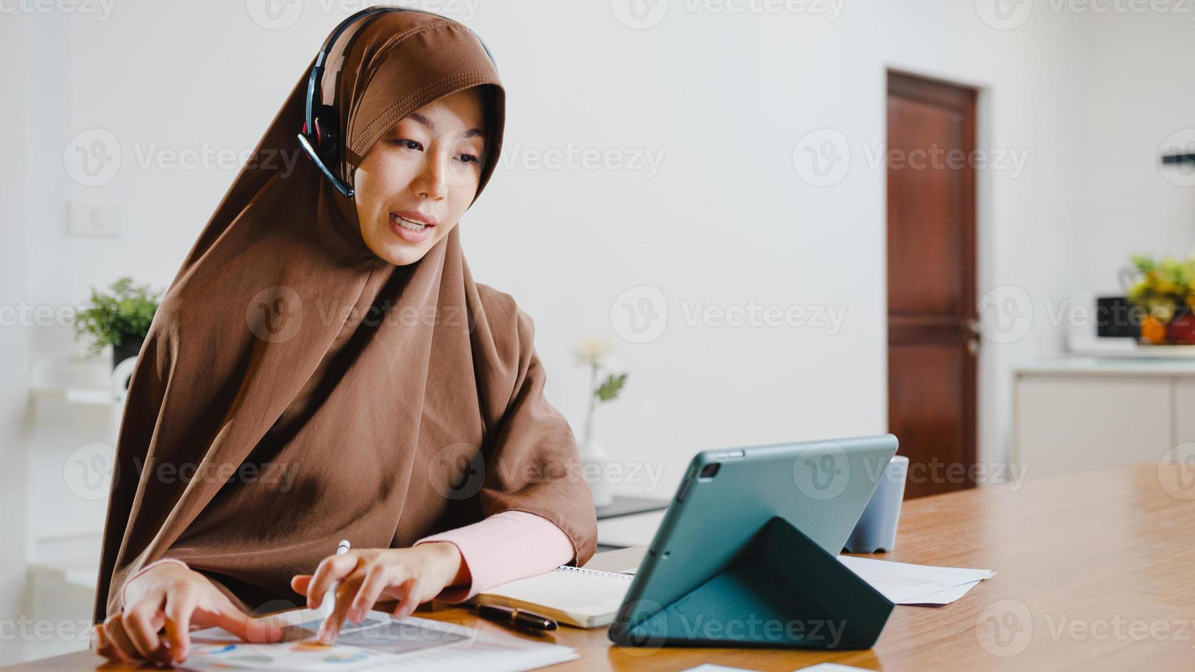 asiatische muslimische dame tragen kopfhörer mit digitaler tablette sprechen mit kollegen über verkaufsbericht in konferenz-videoanrufen, während sie von zu hause in der küche arbeiten. soziale Distanzierung, Quarantäne wegen Corona-Virus. foto