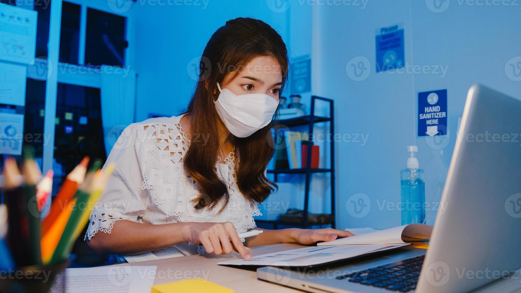 Fröhliche Geschäftsfrau in Asien, die eine medizinische Gesichtsmaske für soziale Distanzierung in einer neuen normalen Situation zur Virenprävention trägt, während sie in der Büronacht den Laptop bei der Arbeit verwendet. Leben und Arbeiten nach dem Coronavirus. foto