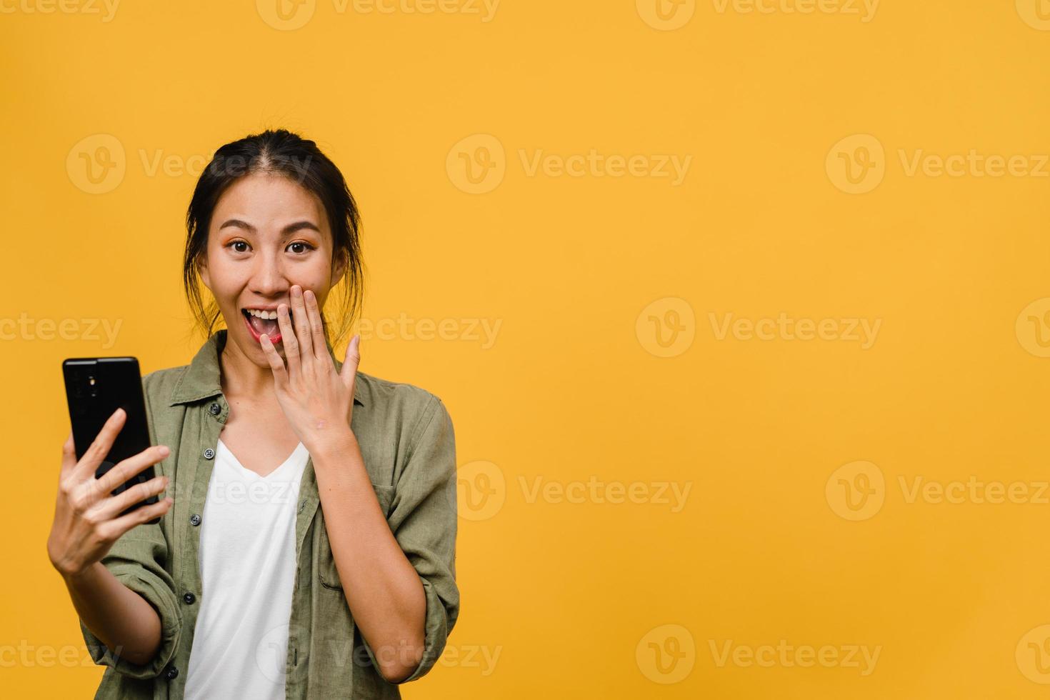 überraschte junge asiatische dame mit handy mit positivem ausdruck, lächelt breit, in legerer kleidung gekleidet und steht einzeln auf gelbem hintergrund. glückliche entzückende frohe frau freut sich über erfolg. foto