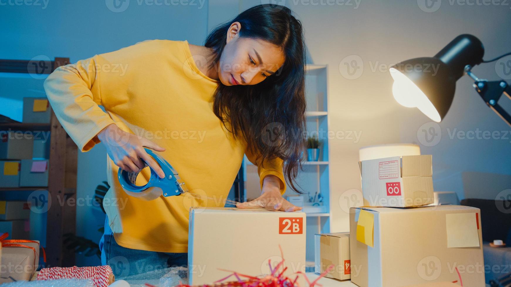 Junge Geschäftsfrau in Asien bereitet Produktverpackungskarton für den Versand an Kundenbestellungen im Home-Office in der Nacht vor. Kleinunternehmer, Online-Marktlieferung, Lifestyle-Freelance-Konzept. foto