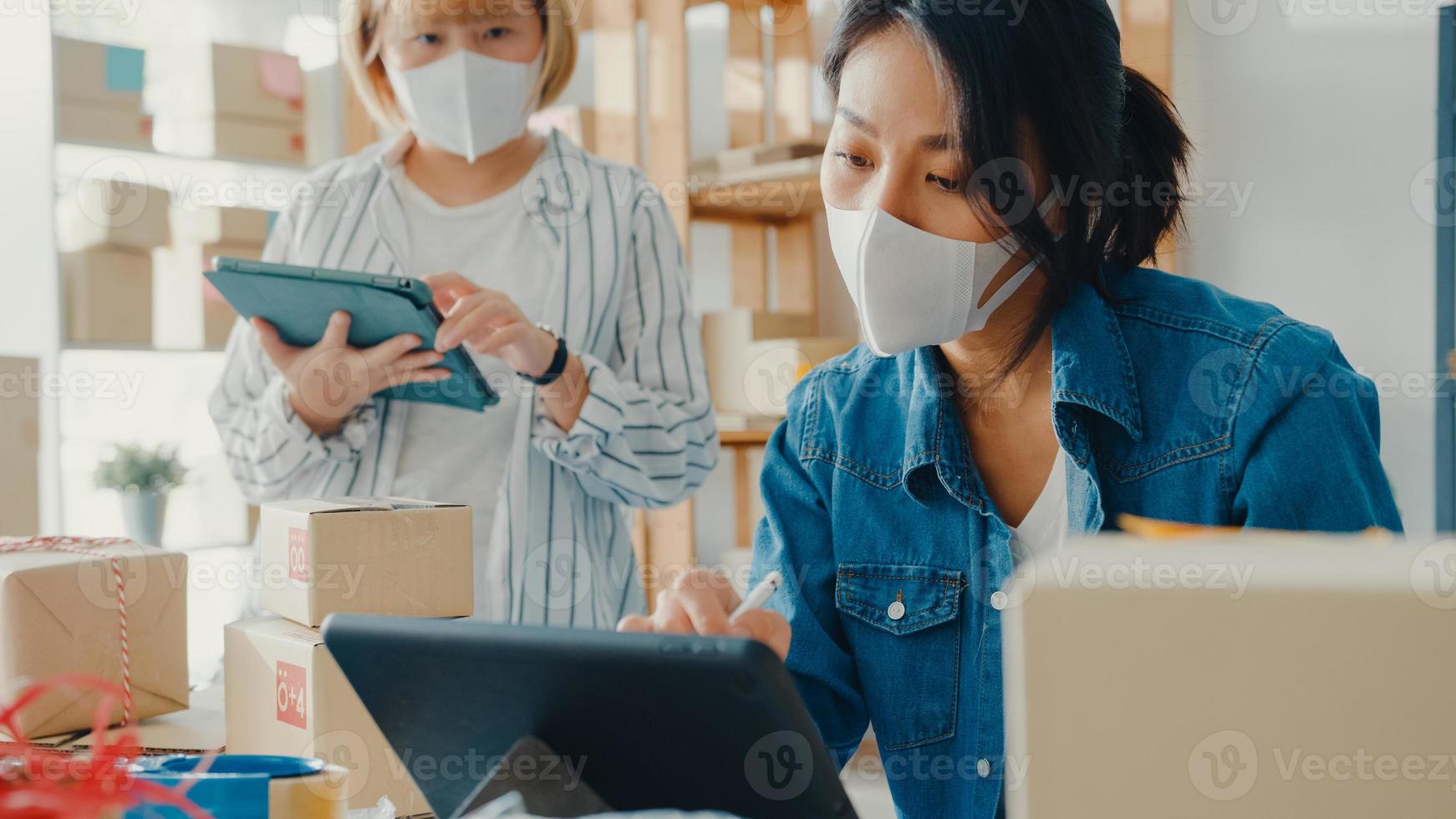 Junge Geschäftsfrauen in Asien tragen Gesichtsmaske Überprüfen Sie die Produktbestellung auf Lager und speichern Sie sie für die Arbeit mit Tablet-Computern im Home Office. Kleinunternehmer, Online-Marktlieferung, Lifestyle-Freelance-Konzept foto