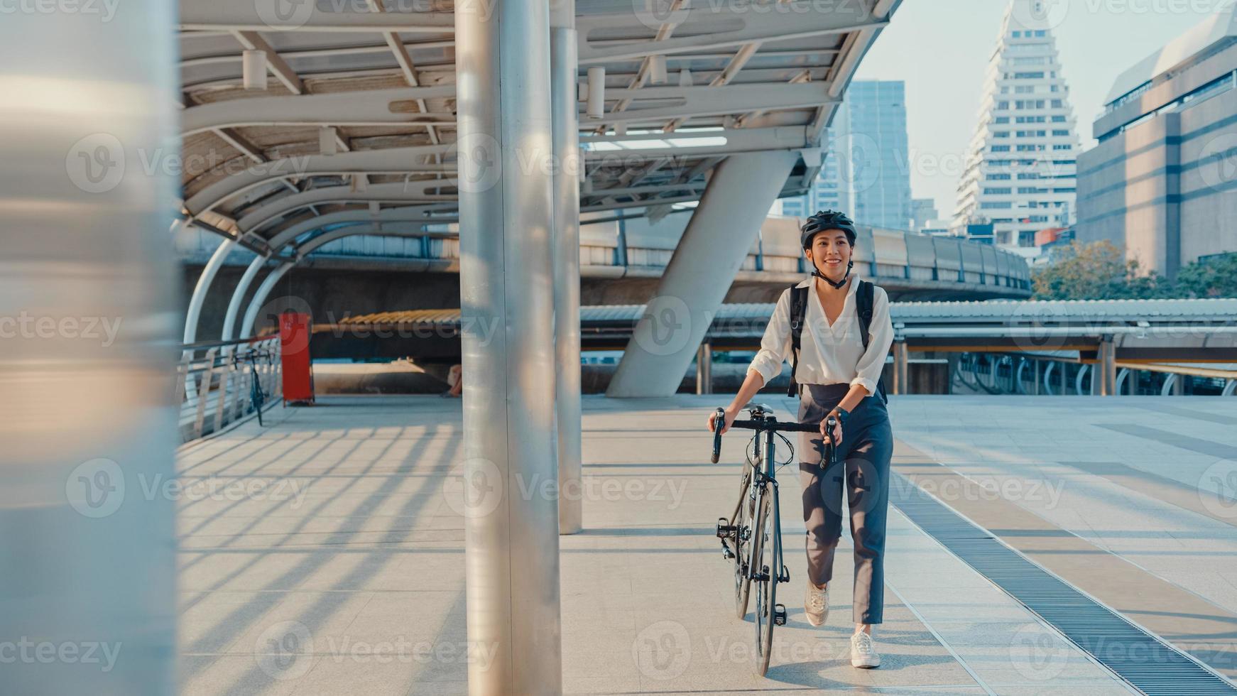 asiatische geschäftsfrau gehen im büro spazieren und lächeln tragen rucksack schauen sich um, nehmen fahrrad auf der straße um das gebäude auf einer stadtstraße. Fahrradpendeln, Pendeln mit dem Fahrrad, Geschäftspendlerkonzept. foto