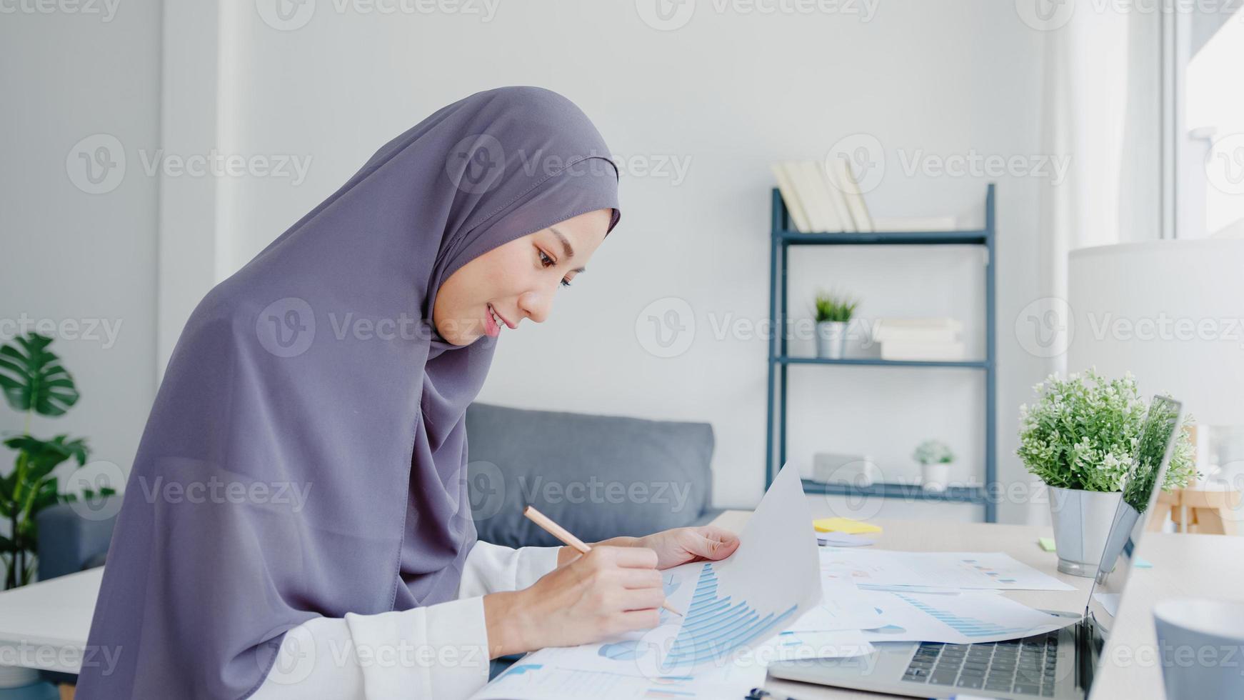 Schöne asiatische muslimische Dame in Kopftuch-Freizeitkleidung mit Laptop im Wohnzimmer im Haus. Fernarbeit von zu Hause aus, neuer normaler Lebensstil, soziale Distanzierung, Quarantäne zur Vorbeugung von Coronaviren. foto