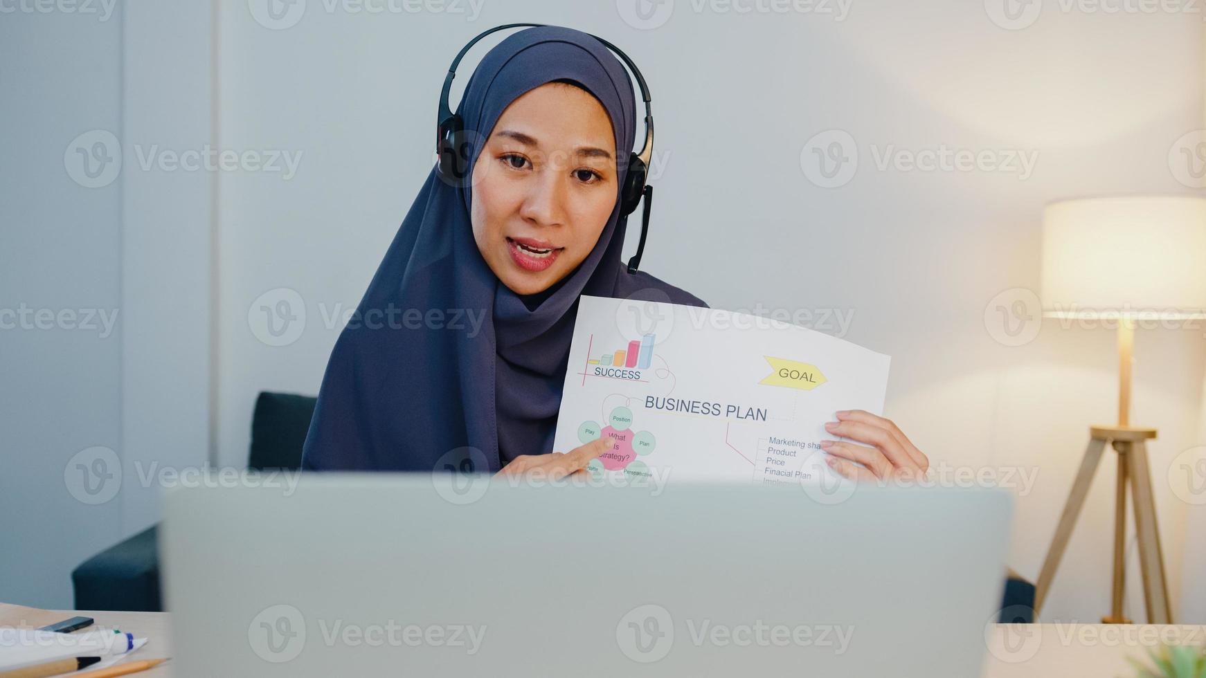 asiatische muslimische dame tragen kopfhörer mit laptop sprechen sie mit kollegen über verkaufsbericht in konferenz-videoanrufen, während sie nachts im heimbüro arbeiten. soziale Distanzierung, Quarantäne wegen Corona-Virus. foto