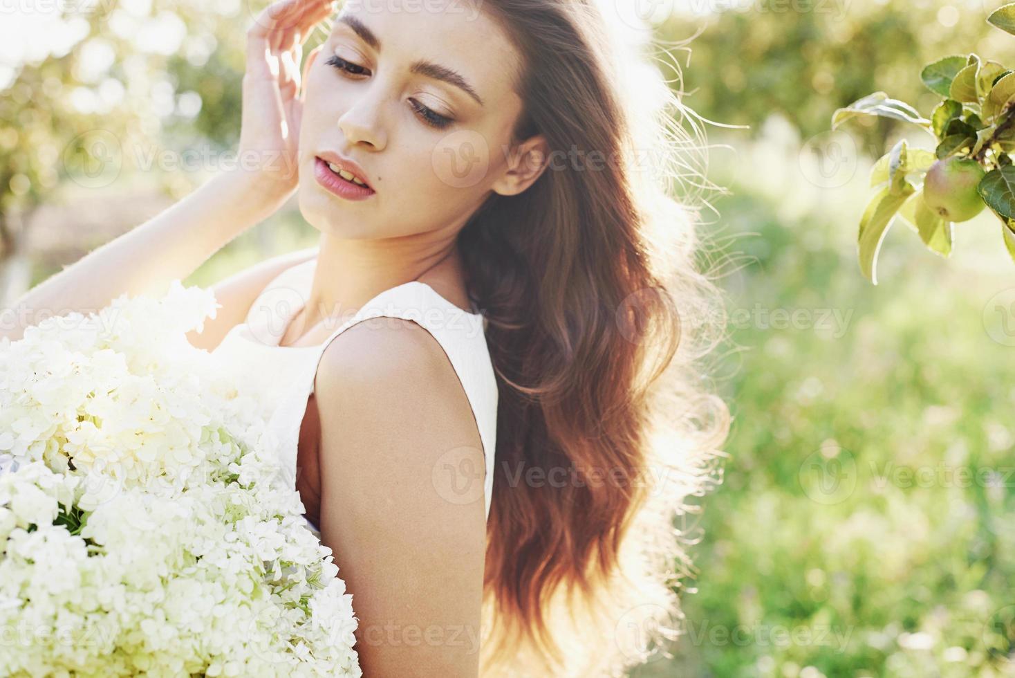 schöne junge frau, die ein elegantes weißes kleid trägt und einen schönen sonnigen nachmittag in einem sommergarten genießt foto