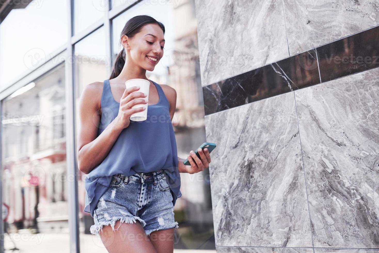 Porträt einer jungen stilvollen Hipster-Frau, die auf der Straße geht, ein süßes trendiges Outfit trägt, heißen Latte-Kaffee trinkt und lächelt foto