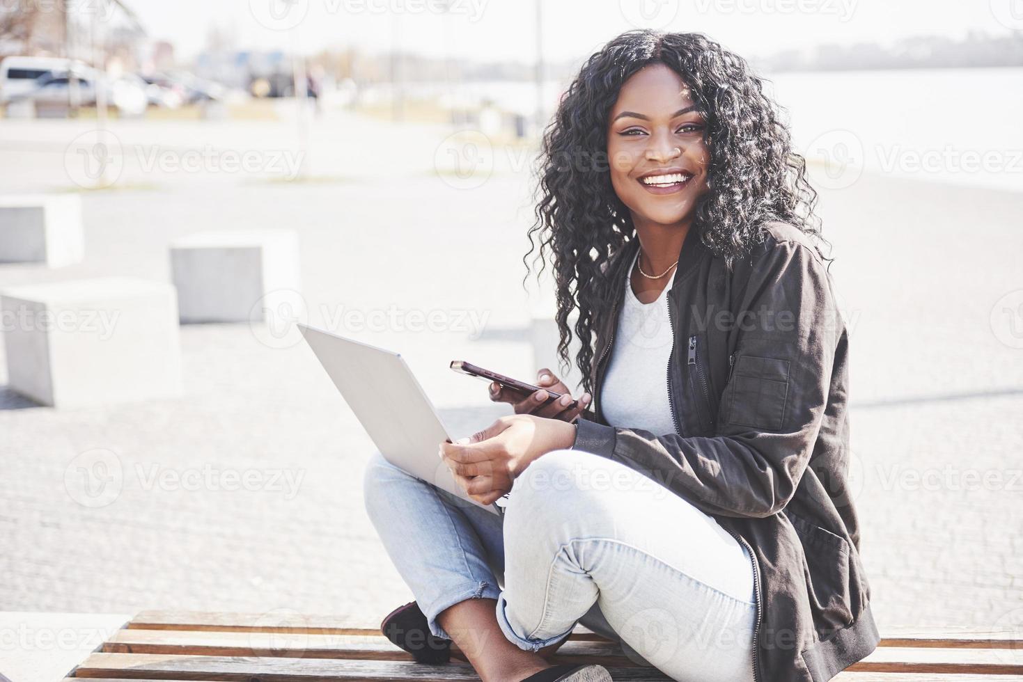 junge Frau auf der Straße, die am Laptop arbeitet und mit dem Handy spricht foto