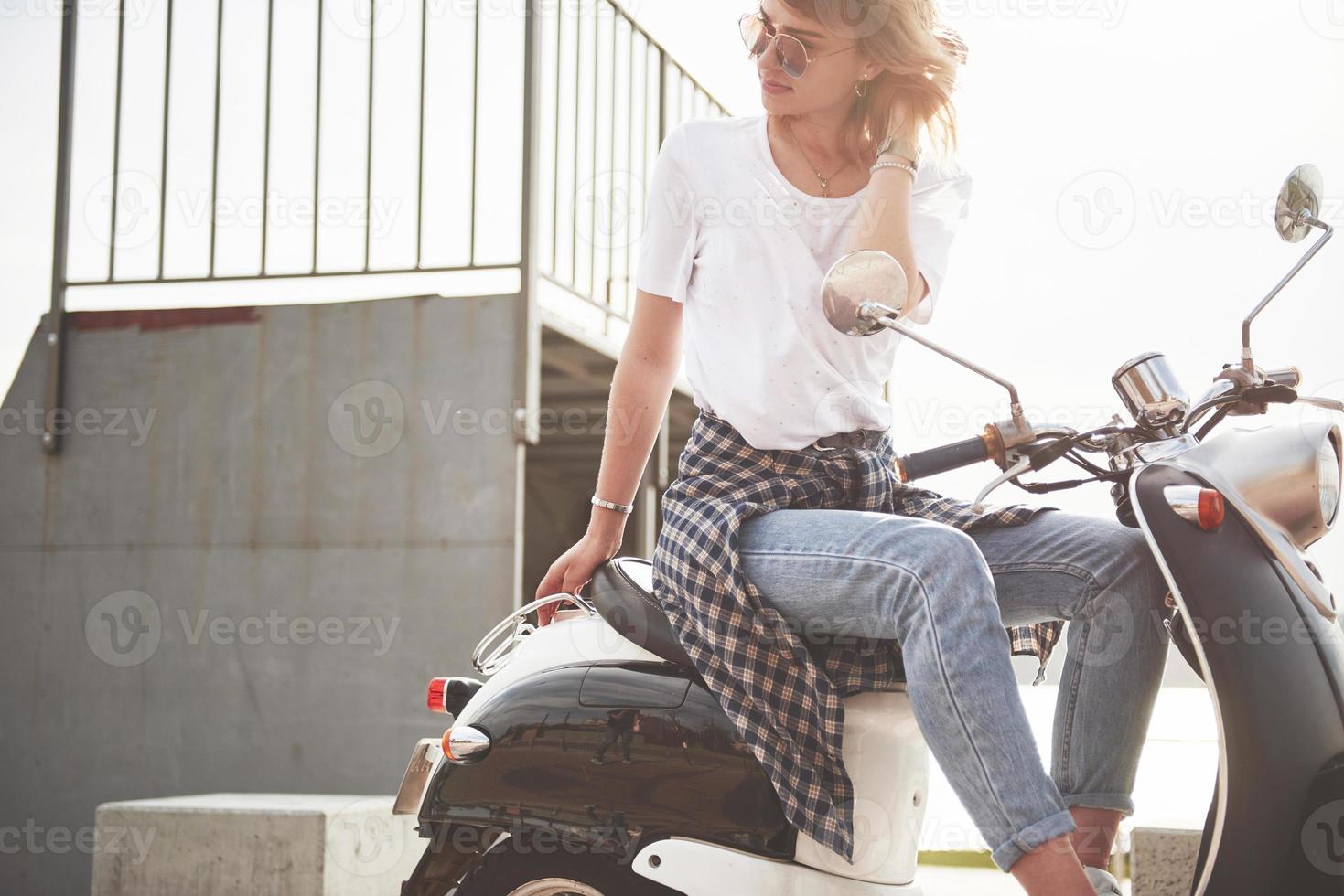 Porträt eines schönen Mädchen-Hipsters, der auf einem schwarzen Retro-Roller sitzt, lächelnd posiert und die warme Frühlingssonne genießt. foto
