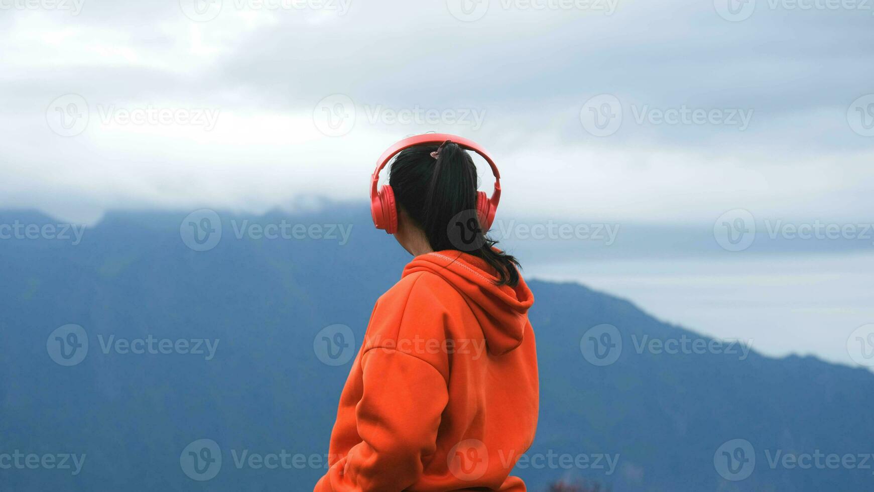 Rückansicht einer jungen Frau, die auf einem ruhigen Hügel steht und morgens Musik über Kopfhörer hört. frau, die einen pullover trägt, der die schönheit der natur genießt, die im winter den berg betrachtet. foto