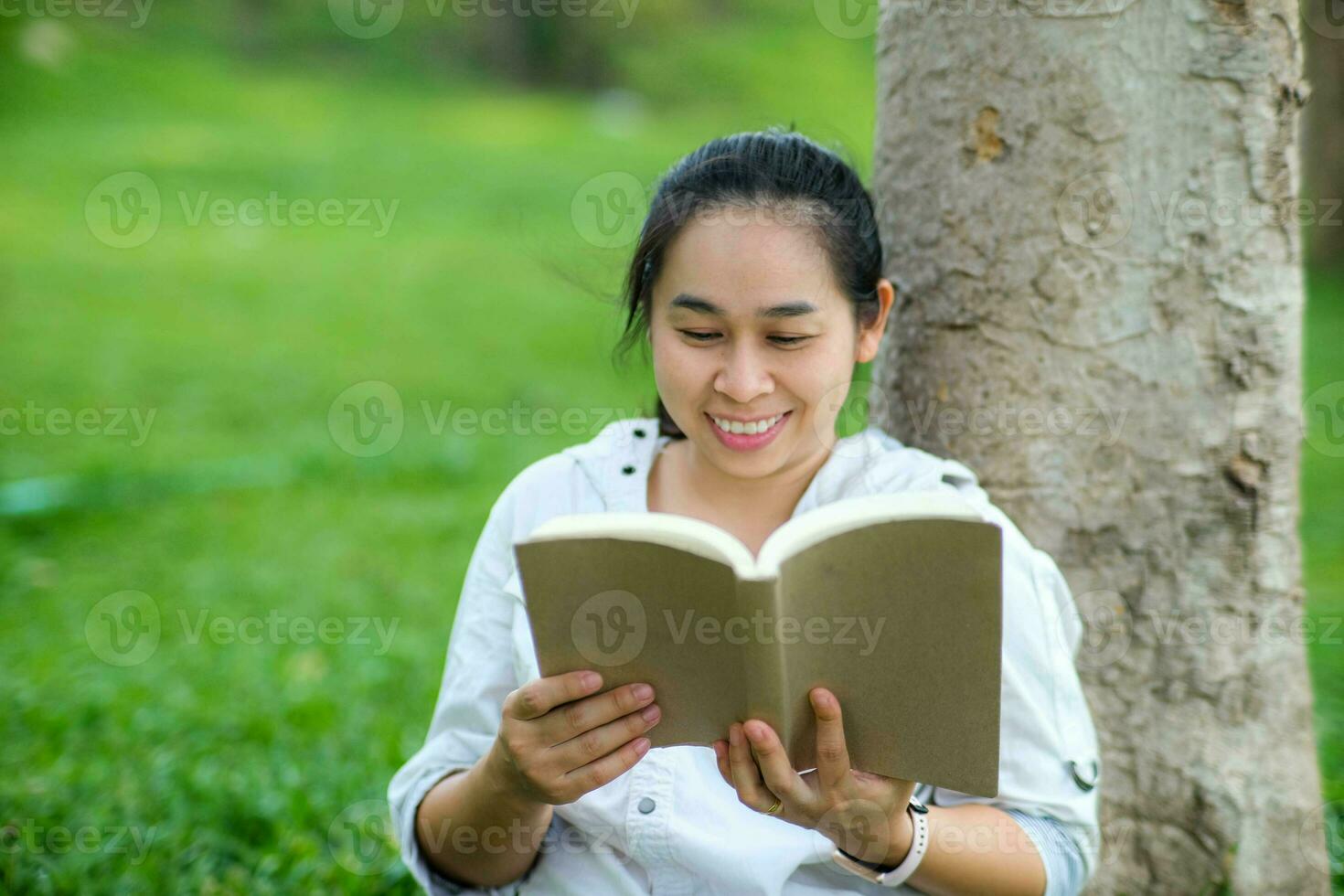 heiter jung Frau im Jacke lesen ein Buch im Sommer- Park. konzentriert Frau Sitzung auf Gras und studieren unter Baum während Urlaub. Bildung Konzept foto