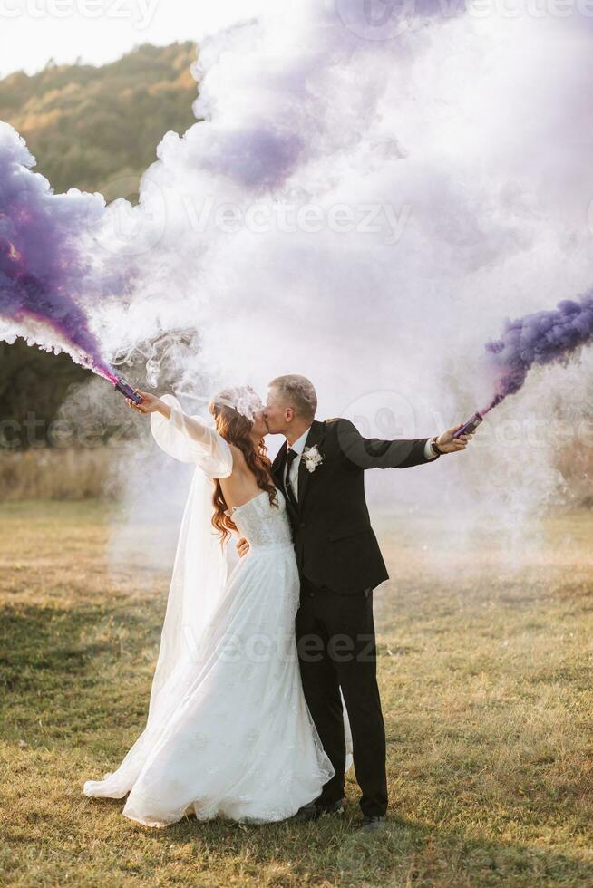 Braut und Bräutigam spielen mit farbig Rauch im lila Hände, Umarmungen und Küsse. Rauch Bomben beim ein Hochzeit. foto