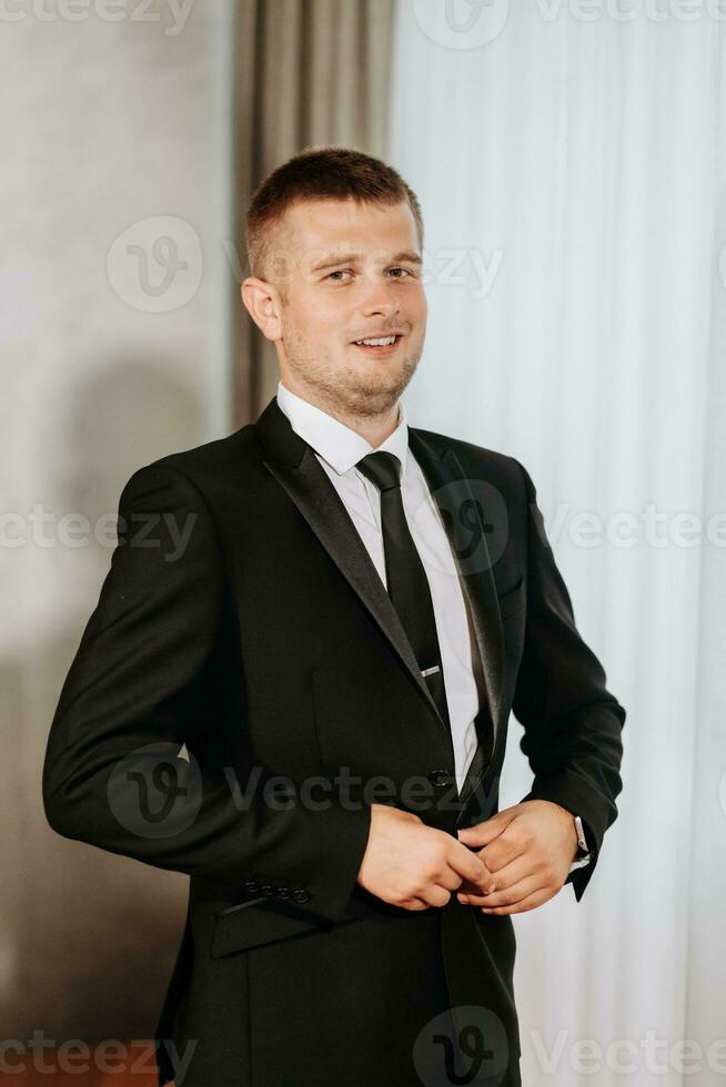 Porträt von ein Erwachsene Mann Knöpfen oben ein schwarz Jacke im seine Zimmer. Vorbereitung von das Bräutigam zum das Hochzeit Zeremonie. foto