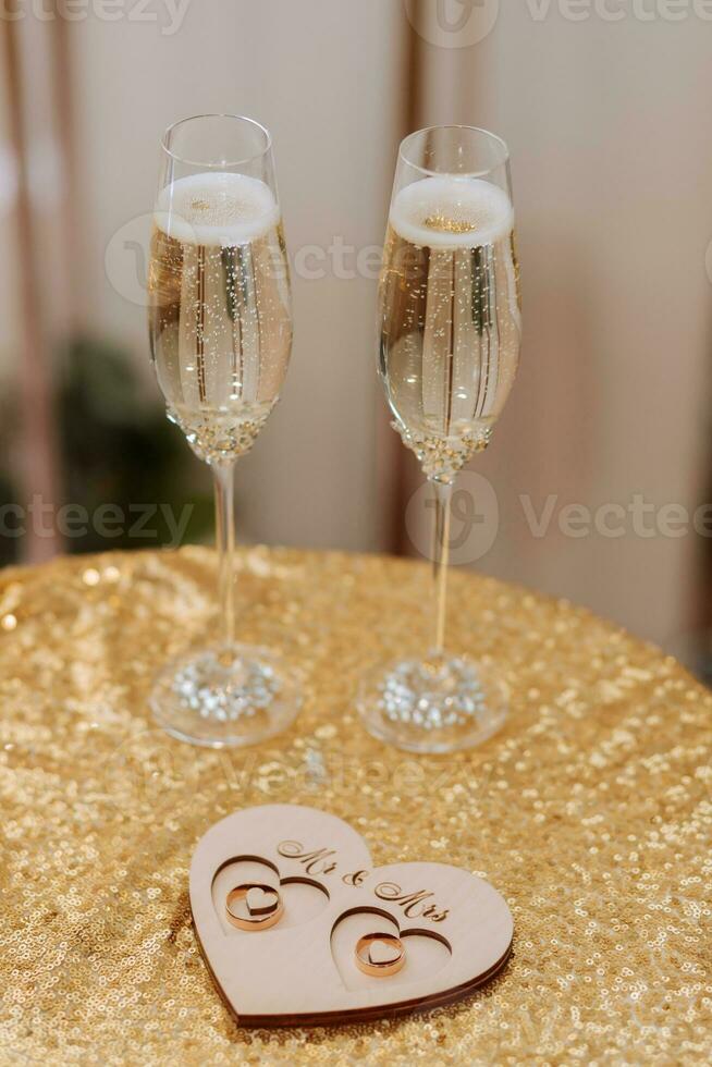 Hochzeit Brille gefüllt mit Champagner auf ein Tabelle bedeckt mit Gold, Nächster zu Gold Hochzeit Ringe auf ein hölzern Stand foto