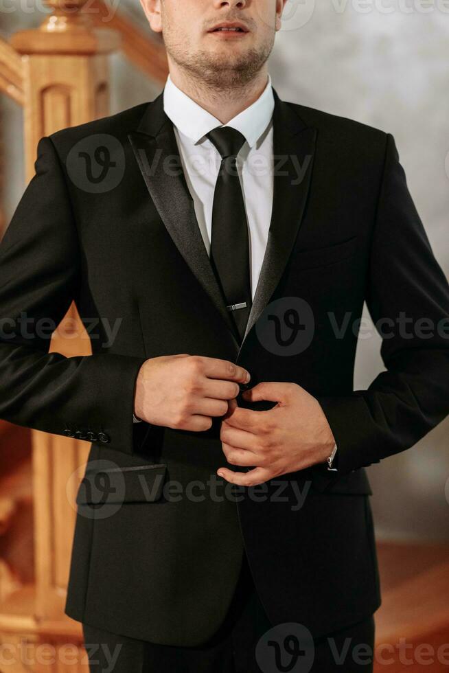 Porträt von ein Erwachsene Mann Knöpfen oben ein schwarz Jacke im seine Zimmer. Vorbereitung von das Bräutigam zum das Hochzeit Zeremonie. foto