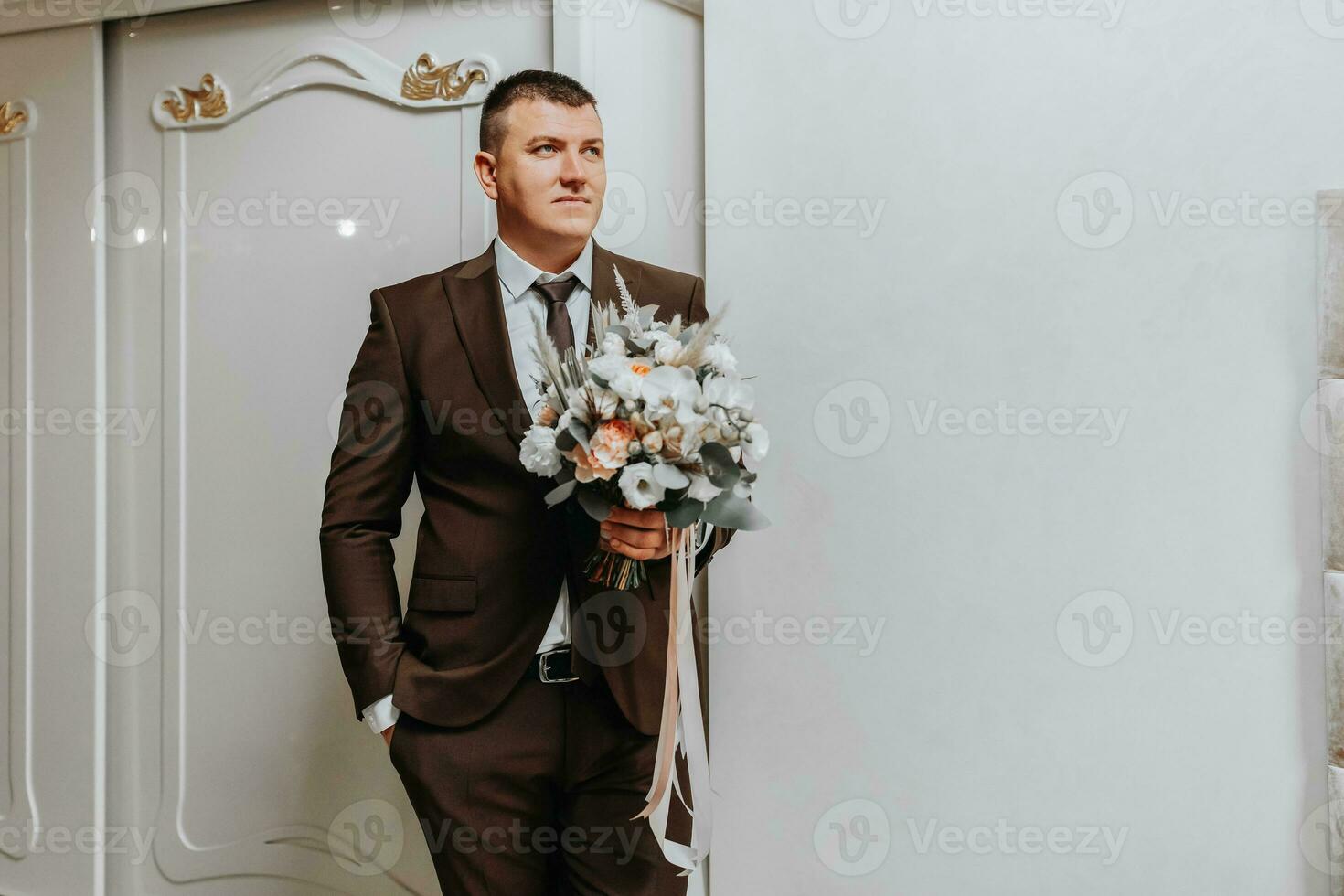 das Bräutigam steht im das Zimmer mit ein Strauß von Blumen im ein klassisch braun passen und Weiß Shirt. Vorbereitung zum das Hochzeit Zeremonie foto