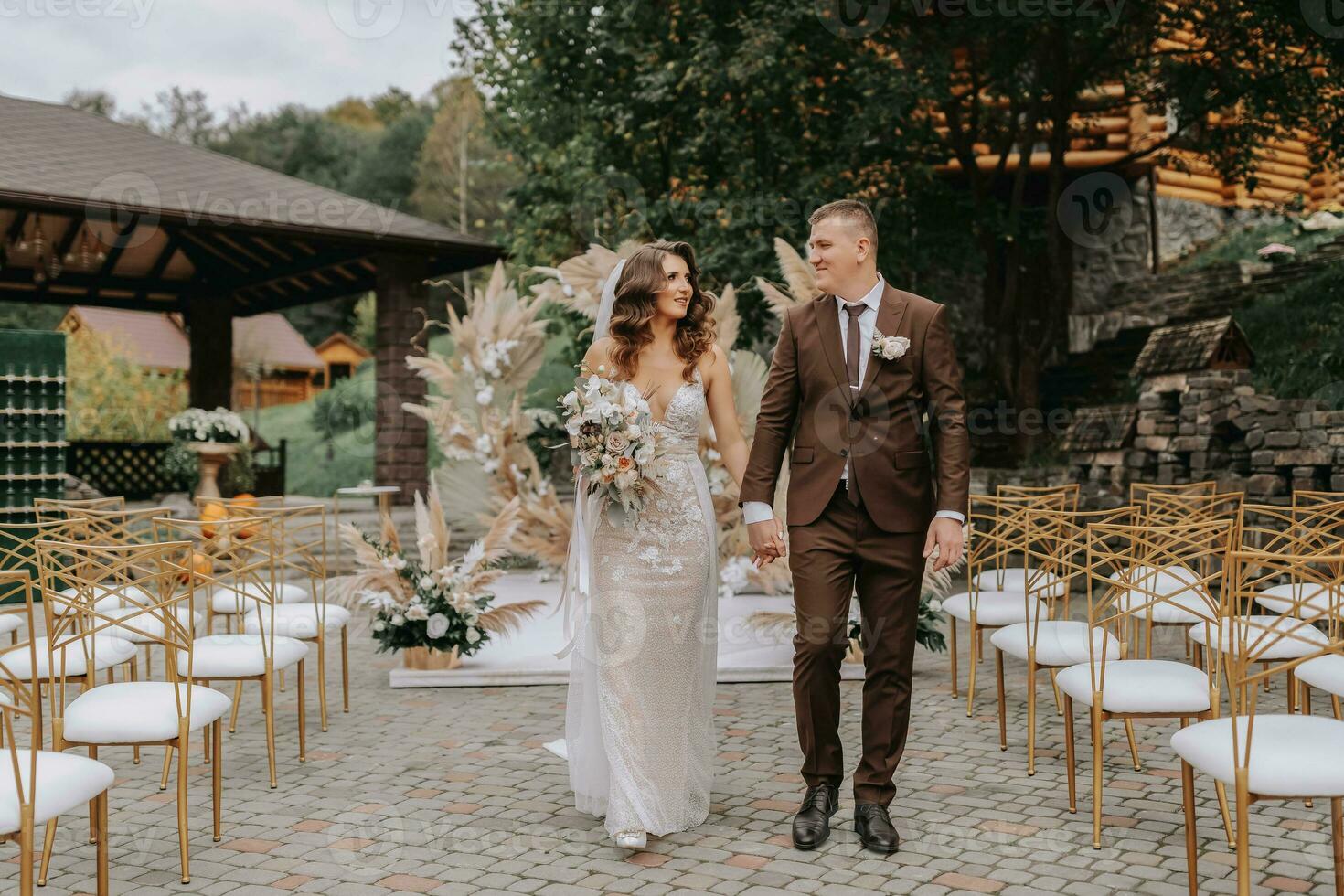 Bräutigam und Braut auf das Hintergrund von das Hochzeit Bogen. Jungvermählten mit ein Hochzeit Strauß, Gehen nach das Hochzeit Zeremonie, dekoriert mit Blumen und getrocknet Blumen im das öffnen Luft. foto