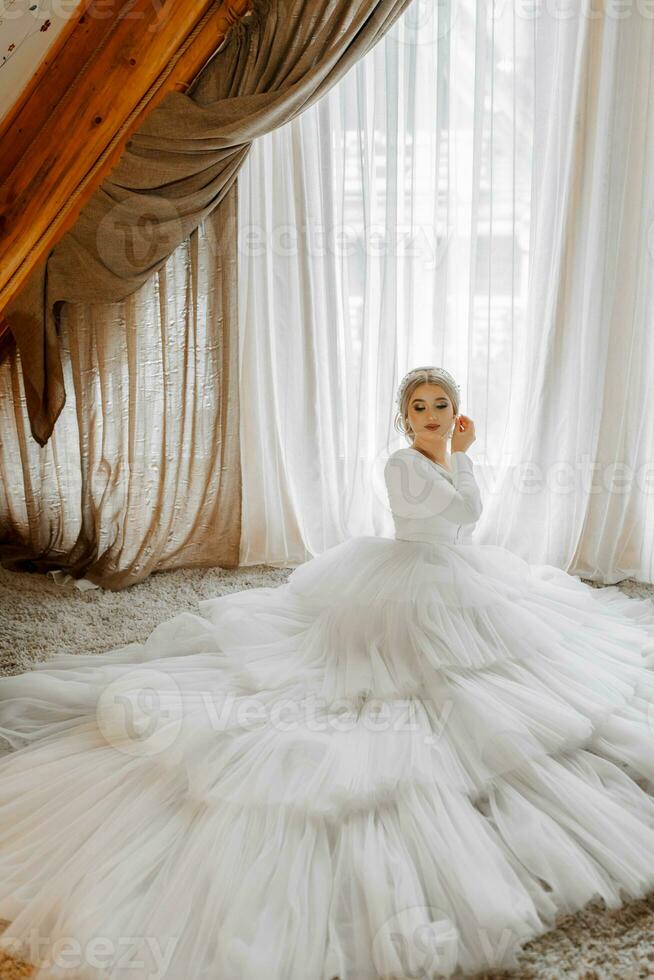 Porträt von ein schön Braut im ein Weiß Hochzeit Kleid mit ein lange Zug im ein Hotel Zimmer. das Braut ist Sitzung auf das Fußboden gegen ein groß Fenster foto