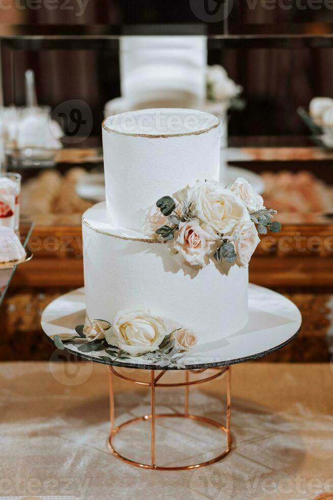 ein Weiß zweistufig Hochzeit Kuchen, dekoriert mit Weiß Blumen und Asche um das Kanten, steht auf ein Stand in der Nähe von ein Spiegel. Hochzeit Nachspeisen. foto