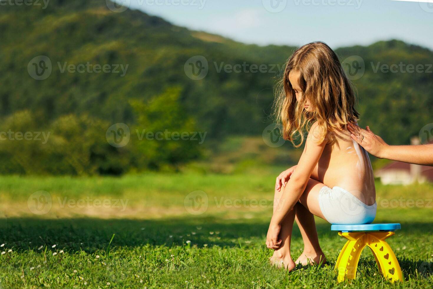 ein Mutter gilt Sonnenschutz zu ihr wenig Tochter zurück Vor das Kind geht zum ein gehen. das Kind ist Sitzung auf ein Stuhl und Grün Gras. kostenlos Raum foto