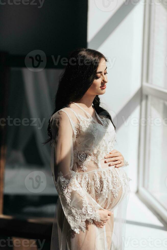 ein schön schwanger Frau im ein Spitze transparent Kleid Umarmungen ihr Bauch mit ihr Hände in der Nähe von das Fenster. Konzept von Schwangerschaft, Mutterschaft, Vorbereitung und warten. das Schönheit von ein Frau während Schwangerschaft foto