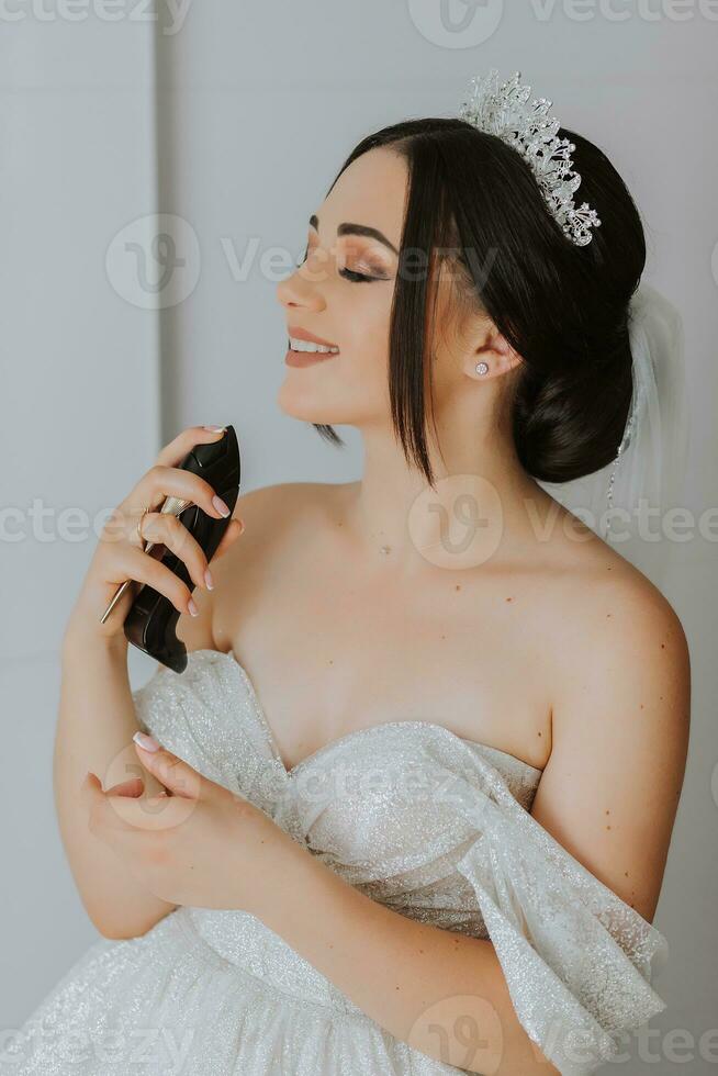 ein Brünette Braut im ein schick Kleid, Frisur und ein Tiara auf ihr Kopf Sprays Parfüm auf ihr Körper im ihr Zimmer foto