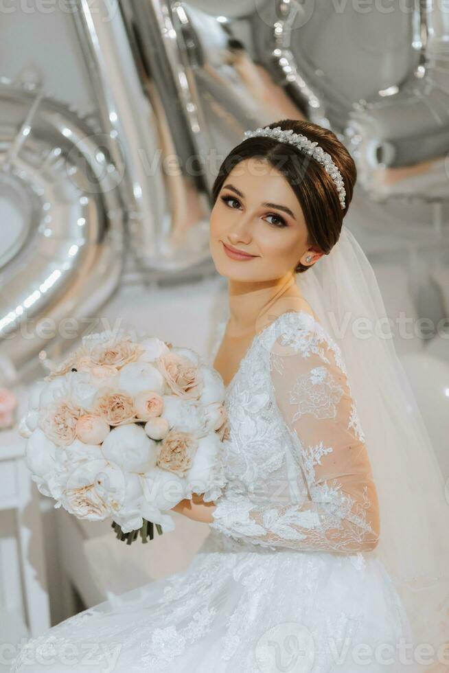 ein schön jung Braut im ein Hochzeit Kleid mit ein Strauß von Pfingstrosen im ihr Hände ist Sitzung auf ein Bett im ein Zimmer. Finale Vorbereitungen zum das Hochzeit. das Braut ist warten zum das Bräutigam. Morgen, Braut. foto