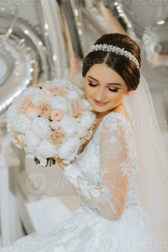 ein schön jung Braut im ein Hochzeit Kleid mit ein Strauß von Pfingstrosen im ihr Hände ist Sitzung auf ein Bett im ein Zimmer. Finale Vorbereitungen zum das Hochzeit. das Braut ist warten zum das Bräutigam. Morgen, Braut. foto