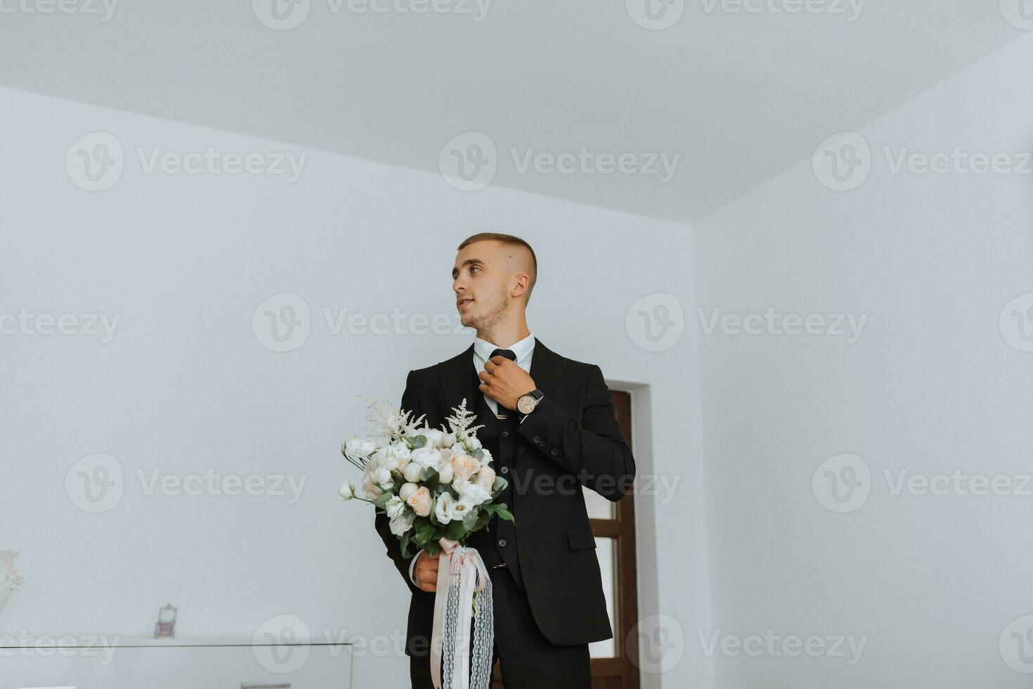 Porträt. ein Mann im ein Weiß Shirt, schwarz passen posiert im ein Weiß Zimmer, halten ein Strauß. ein stilvoll betrachten. Herren Stil. Mode. Geschäft foto