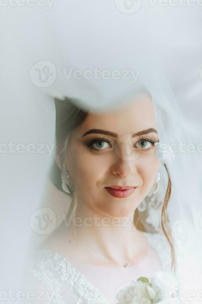 das Braut ist gekleidet im ein elegant Kleid, bedeckt mit ein Schleier, posieren und halten ein Ansteckblume. Hochzeit Foto, Morgen von das Braut foto