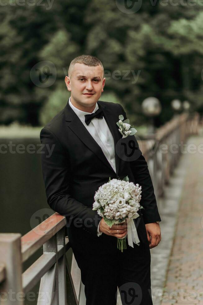 Hochzeit Porträt. ein Mann im ein schwarz passen steht auf ein Brücke mit ein Strauß und sieht aus weg. Verlobter. männlich, Geschäft Stil foto