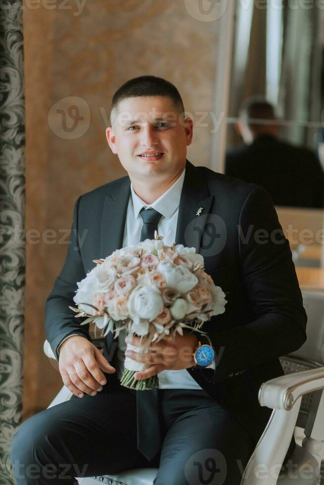 ein stilvoll Bräutigam mit ein Strauß von Blumen ist Sitzung auf ein Stuhl im ein teuer Hotel Zimmer. Bräutigam Morgen. das Bräutigam ist bekommen bereit im das Morgen Vor das Hochzeit Zeremonie foto