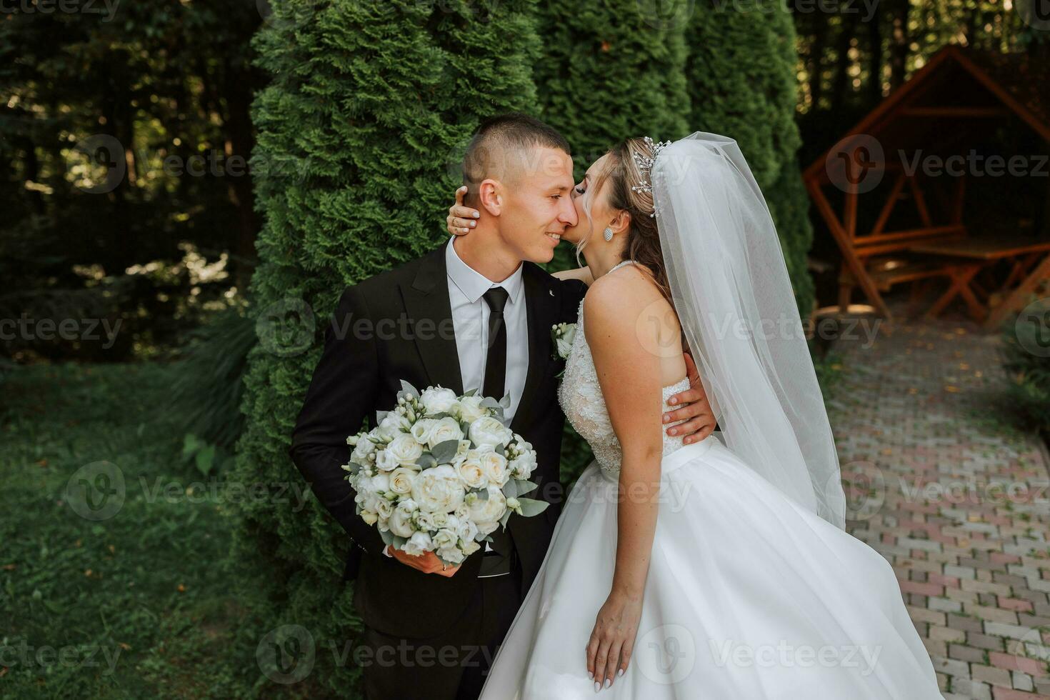 ein stilvoll Bräutigam im ein schwarz passen und ein süß Braut im ein Weiß Kleid mit ein lange Schleier sind umarmen im ein Park. Hochzeit Porträt von lächelnd und glücklich Jungvermählten. foto