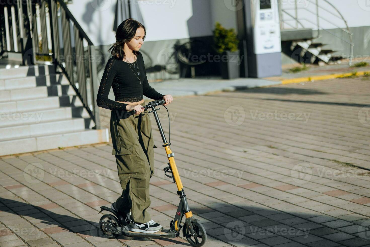 draussen Porträt von jung Teenager Brünette Mädchen mit lange Haar Fahren Roller auf Stadt Straße foto