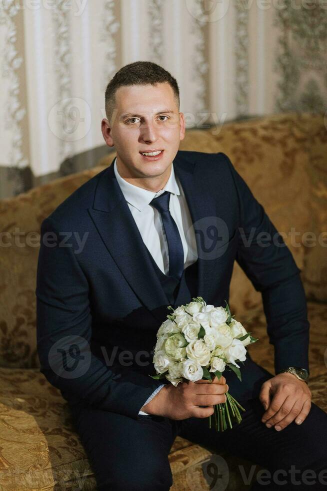 das elegant Bräutigam stellen auf ein Jacke im seine Zimmer. Einzelheiten von das Hochzeit Morgen. Einzelheiten Schönheit und Mode. Bräutigam mit ein Strauß von Blumen foto