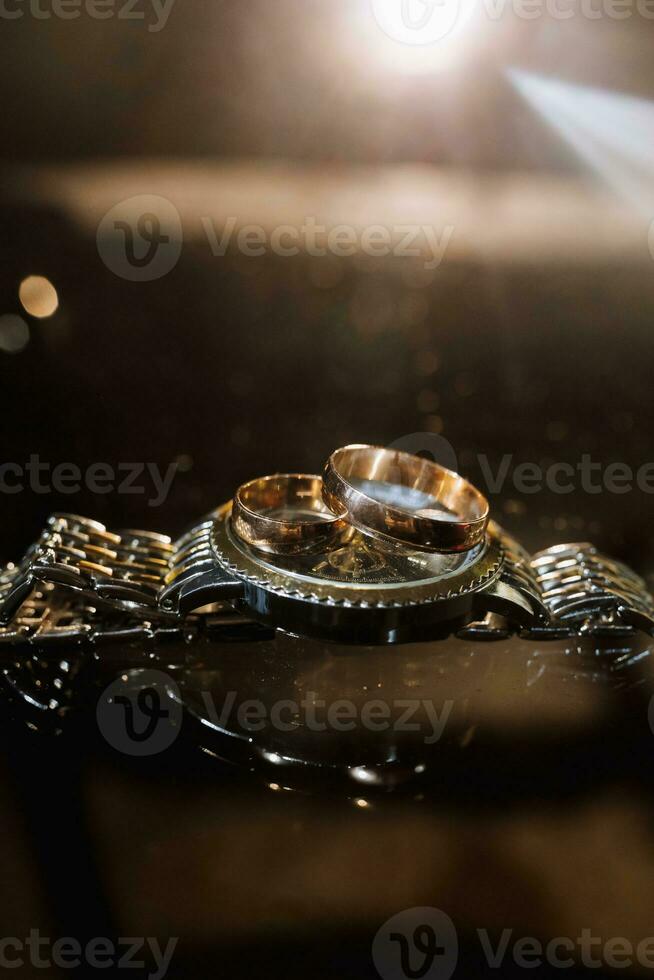 Zubehör zum das Bräutigam Hochzeit Tag. Gold Ringe auf ein Uhr auf ein dunkel Hintergrund mit Hintergrundbeleuchtung. Herren Mode foto