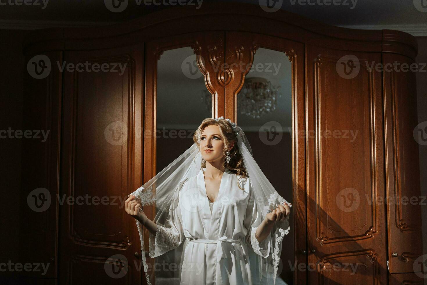 Vorbereitung zum das Hochzeit. schön jung Braut im ein Weiß Kleid im das Zimmer. ein luxuriös Modell- in der Nähe von ein Spiegel mit ein Schleier und ein Tiara auf ihr Kopf. Morgen von das Braut foto