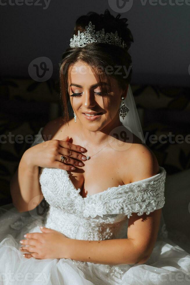 das Braut ist Sitzung im ein Hochzeit Kleid durch das Fenster unter das Sonne Strahlen foto