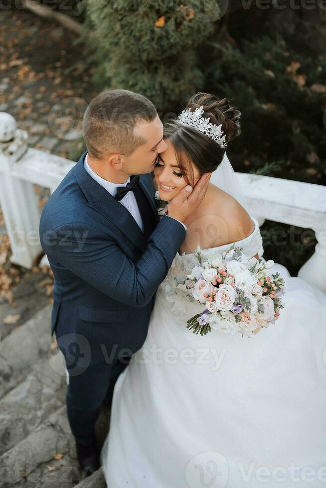 Porträt von glücklich Hochzeit Paar, Braut und Bräutigam im Herbst Wald, Park posieren in der Nähe von Stein Stufen. ein Mann im ein Anzug, ein Mädchen im ein Hochzeit Kleid. Bräutigam Küsse seine Freundin. Foto von über