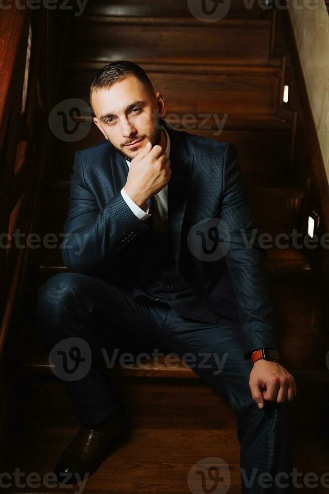 elegant Bräutigam mit ein Bart im ein schön Blau Jacke und Weiß Shirt. Geschäftsmann im klassisch Kleidung. Brünette Mann sitzt auf hölzern Stufen. Studio Porträt foto