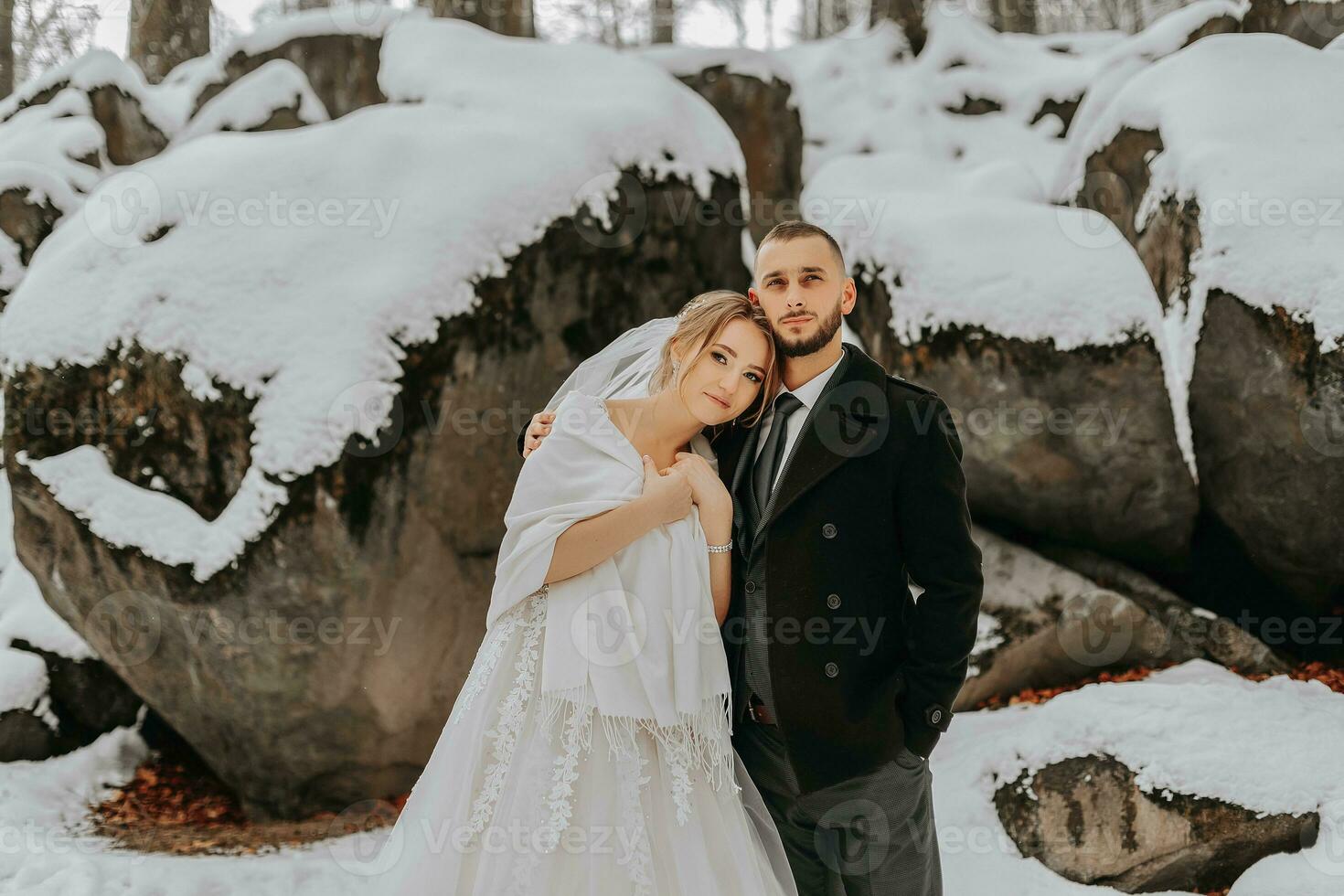 Hochzeit im Winter. das Braut und Bräutigam sind Stehen gegen das Hintergrund von schneebedeckt Felsen. das Braut im ein Weiß Kleid und Weiß Poncho. Bräutigam im ein schwarz Mantel foto