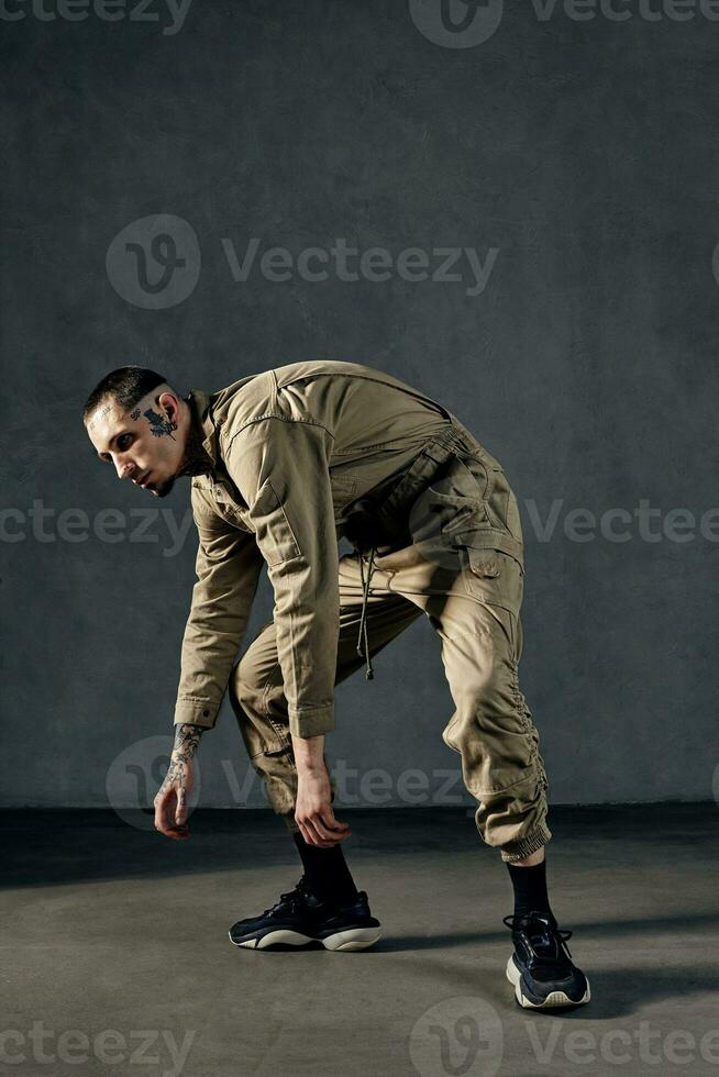 stark Mann mit tätowiert Körper und Gesicht, Ohrringe, Bart. gekleidet im khaki Overall und schwarz Turnschuhe. Tanzen auf grau Hintergrund. Tanzsaal, HipHop foto