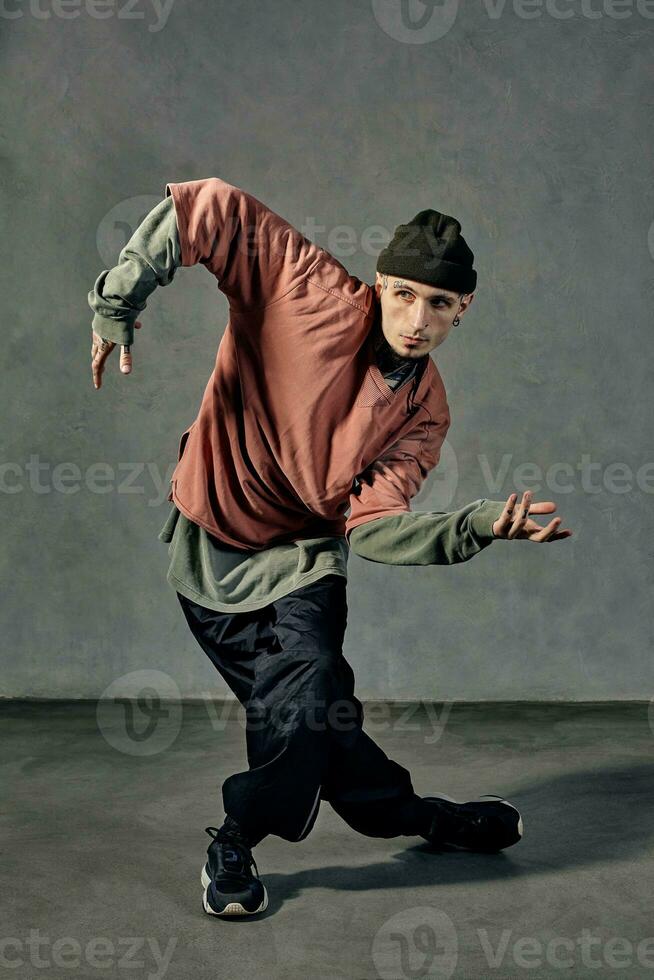 gut aussehend Künstler mit tätowiert Körper, Bart. gekleidet im Hut, beiläufig Kleider und schwarz Turnschuhe. Tanzen auf grau Hintergrund. Tanzsaal, HipHop foto