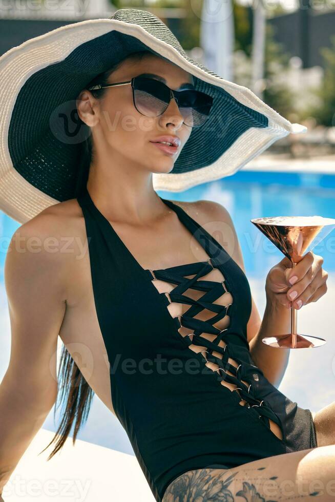 Porträt von ein weiblich haben sich ausruhen und posieren Sitzung in der Nähe von ein Schwimmen Schwimmbad. gekleidet im ein schwarz Badeanzug, Hut und Sonnenbrille. foto