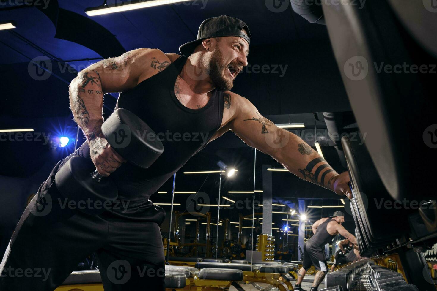 tätowiert, bärtig muskulös Mann im schwarz kurze Hose, Weste, Deckel. ausüben mit Hanteln zum Ausbildung seine Trizeps dunkel Fitnessstudio. Sport, Fitness. schließen oben foto