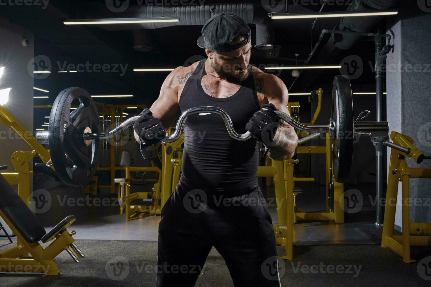 bärtig Mann im schwarz Sport Handschuhe, kurze Hose, Weste und Deckel. Heben ein Hantel, Ausbildung seine Muskeln, Stehen im dunkel Fitnessstudio mit Gelb Ausrüstung. schließen oben foto