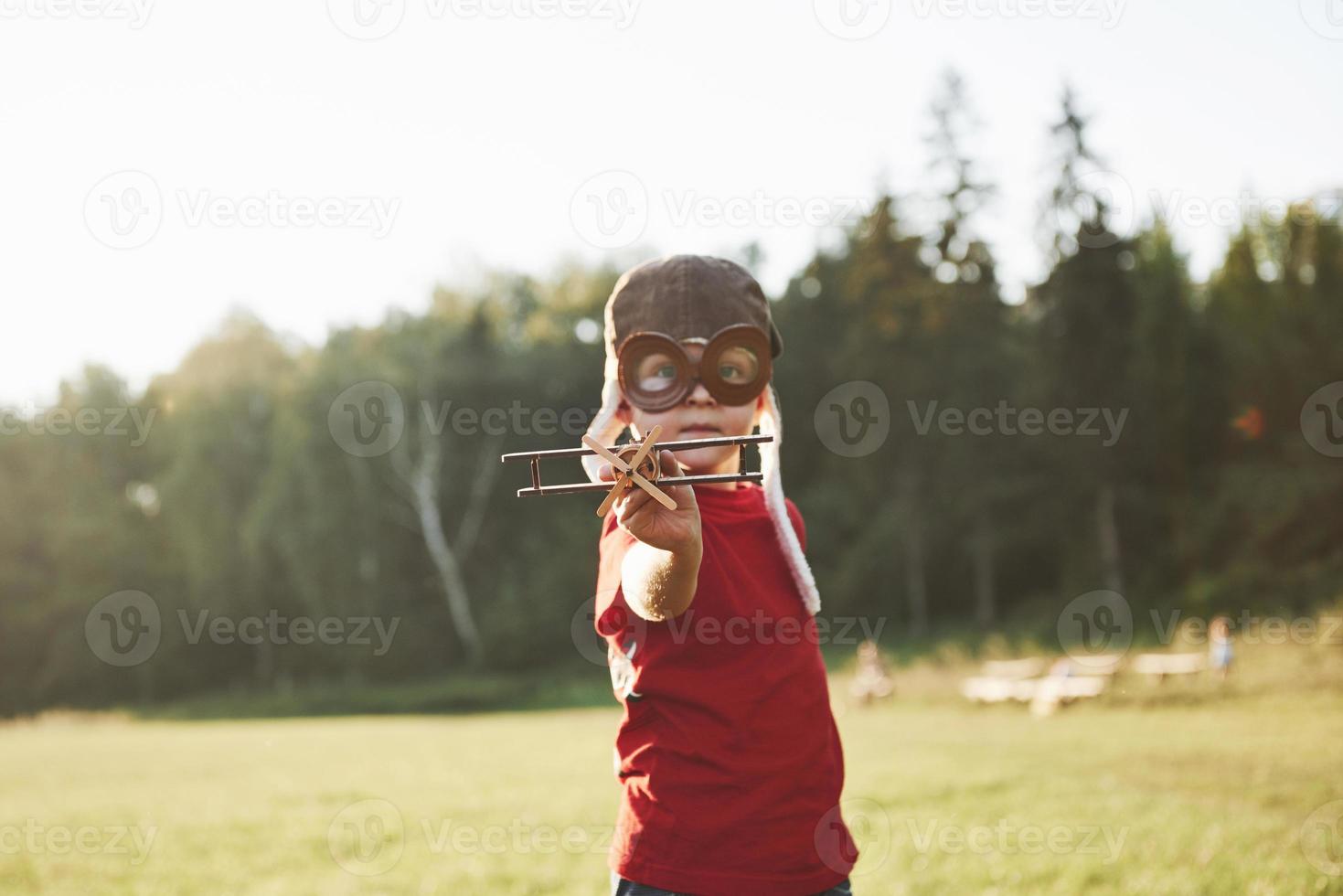 glückliches Kind im Pilotenhelm, das mit einem hölzernen Spielzeugflugzeug spielt und davon träumt, fliegen zu werden foto